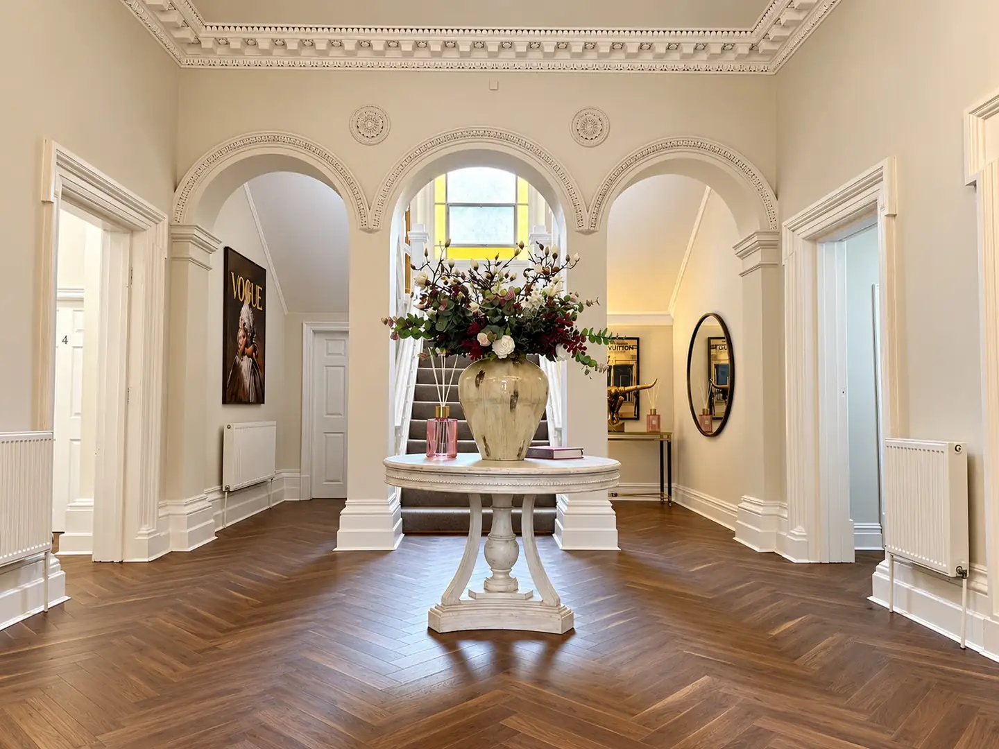 Lobby with a round pedestal table and floral urn in front of three arches and a staircase